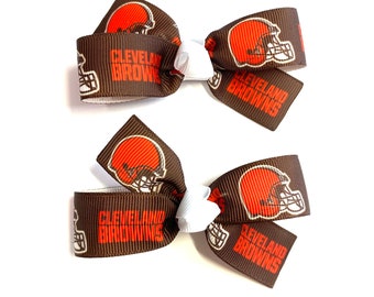 May include: Two brown hair bows with the Cleveland Browns logo. Each bow features the team's helmet in orange and white, with the words "Cleveland Browns" printed in orange. The bows are tied with a white ribbon.