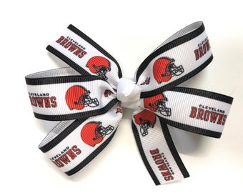 May include: A white ribbon bow featuring the words "Cleveland Browns" in brown and the image of a brown football helmet. The ribbon has black trim.