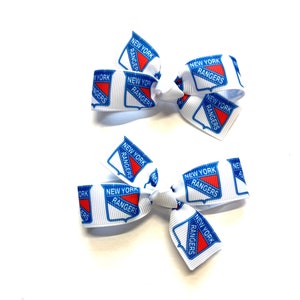 May include: Two white hair bows with a repeating pattern of the New York Rangers logo in red, white, and blue. The bows are made of a ribbon material and are tied in a bow shape. The bows are on a white background.