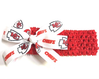 May include: A red crocheted headband with a white bow featuring the Kansas City Chiefs logo and the word "CHIEFS" in red. The bow is centered on the headband, creating a decorative accent.