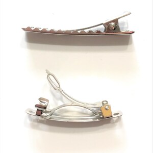 May include: Two silver-colored metal hair clips. One is a crocodile clip with a toothed edge, and the other is a barrette clip with a curved design. Both clips are on a white background.
