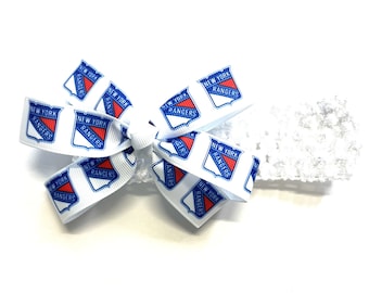May include: A white headband with a decorative bow featuring the New York Rangers logo. The bow is made of white ribbon printed with the team's logo in red, white, and blue. The headband is white.