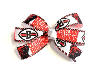 May include: A white, red, and black hair bow featuring the Cleveland Browns logo and team name. The bow has a bulldog graphic and the text "Cleveland Browns" printed on the ribbon. The bow is a fan accessory.