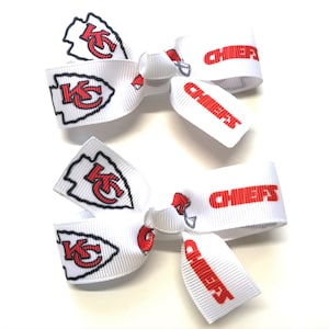 May include: Two white hair bows featuring the Kansas City Chiefs logo and the word "CHIEFS" in red. The bows have a classic bow shape, with the team's arrowhead logo prominently displayed.