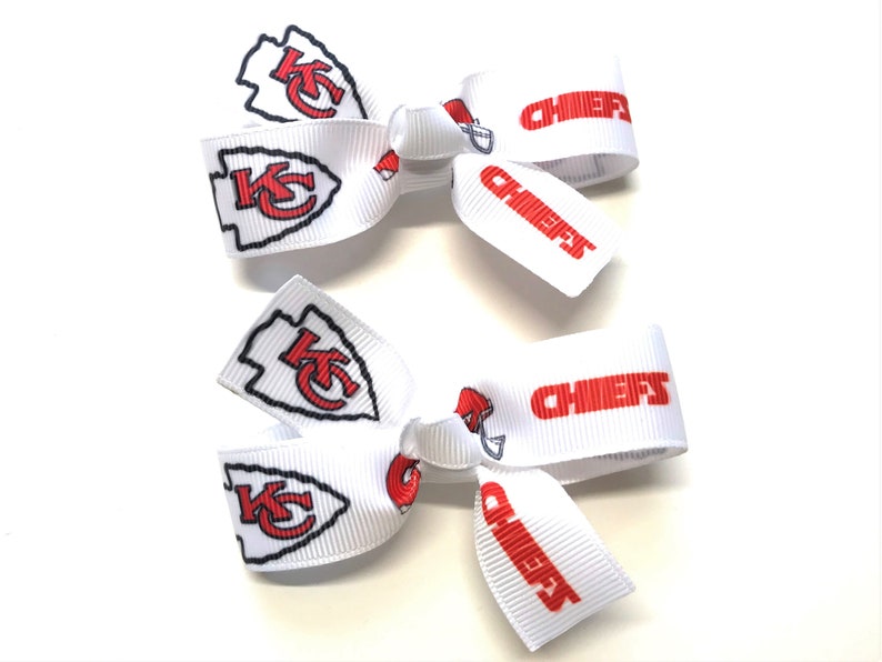 May include: Two white hair bows featuring the Kansas City Chiefs logo and the word "CHIEFS" in red. The bows have a classic bow shape, with the team's arrowhead logo prominently displayed.