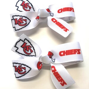 May include: Two white hair bows with red and black Kansas City Chiefs logos and the word "CHIEFS" printed in red. The bows are made of a ribbed fabric and are displayed on a white surface.