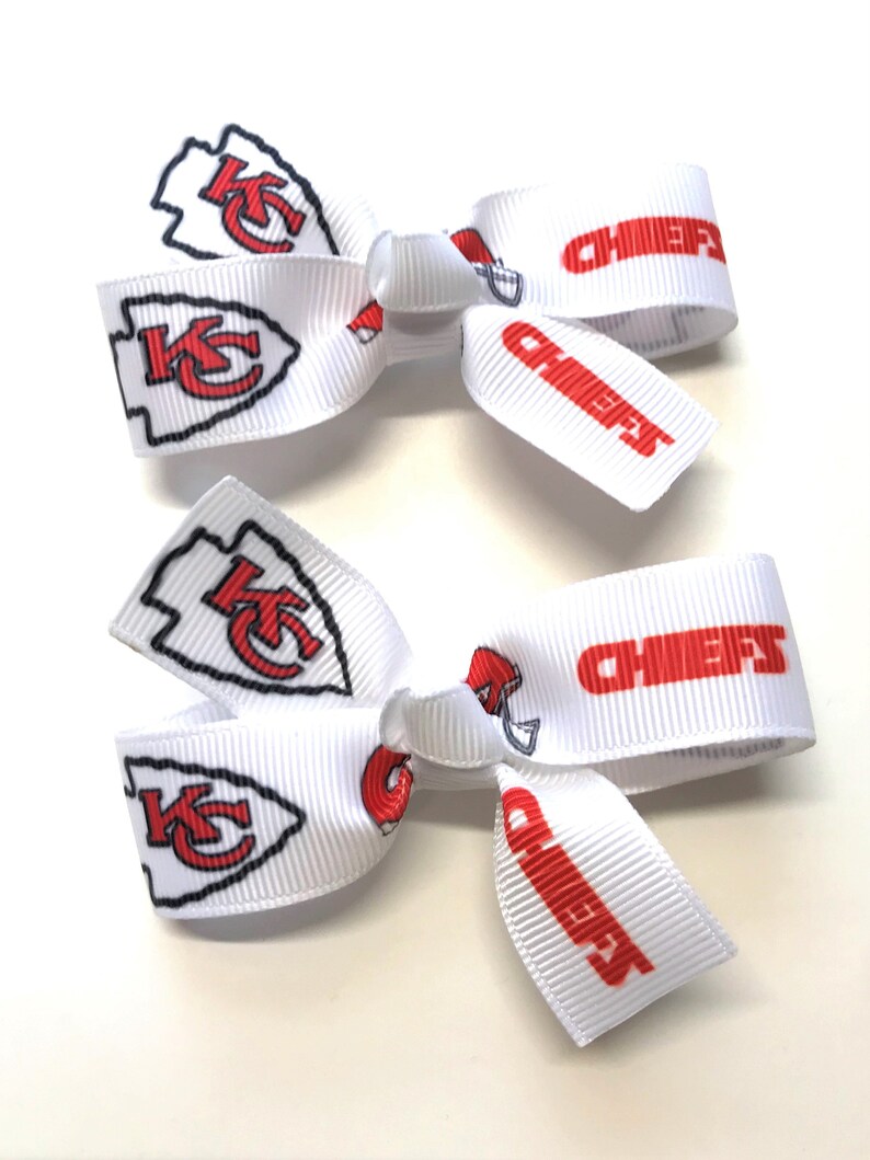 May include: Two white hair bows with red and black Kansas City Chiefs logos and the word "CHIEFS" printed in red. The bows are made of a ribbed fabric and are displayed on a white surface.