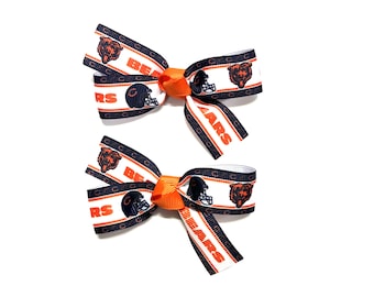 May include: Two Chicago Bears themed hair bows. The bows are white with navy blue and orange accents. The bows feature the team's logo, a football helmet, and the word "BEARS".