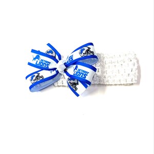 May include: A white crocheted headband with a blue and white bow. The bow features the words "DETROIT LIONS" and a football helmet graphic. The headband is a sports-themed accessory.