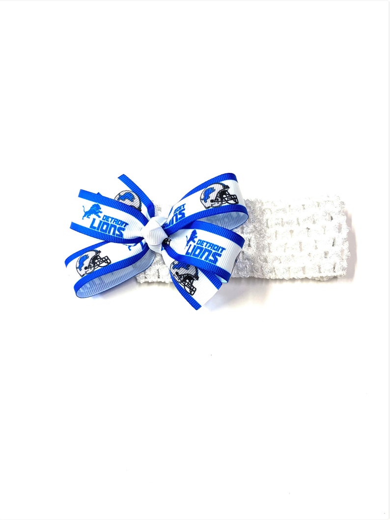 May include: A white crocheted headband with a blue and white bow. The bow features the words "DETROIT LIONS" and a football helmet graphic. The headband is a sports-themed accessory.