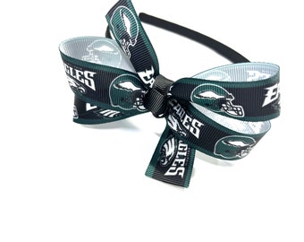 May include: A black headband with a large bow made from ribbon featuring the Philadelphia Eagles logo and helmet design. The bow is green, black, and white, with the team name and logo prominently displayed.