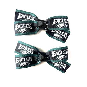 May include: Two Philadelphia Eagles themed hair bows. Each bow is made of black and teal ribbon with the word "EAGLES" and the team logo printed on the ribbon. The bows are arranged on a white background.