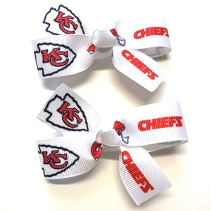 May include: Two white hair bows with the Kansas City Chiefs logo and the word "CHIEFS" in red. The bows feature the team's arrowhead logo and helmet design. Perfect for game day or showing team spirit.