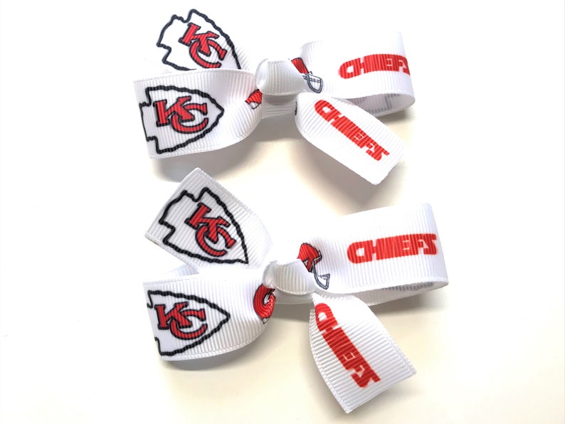 May include: Two white hair bows with the Kansas City Chiefs logo and the word "CHIEFS" in red. The bows feature the team's arrowhead logo and helmet design. Perfect for game day or showing team spirit.