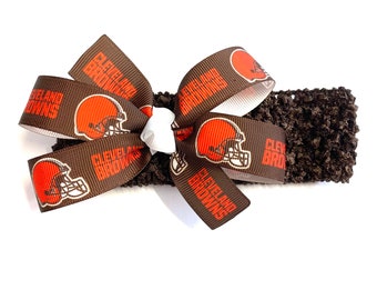 May include: A brown and orange Cleveland Browns bow on a dark brown crocheted headband. The bow features the team's logo and name printed on the ribbon. The headband is a dark brown color.