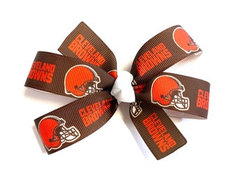 May include: A brown and orange hair bow featuring the Cleveland Browns football team logo. The bow is made of ribbon with the team's helmet and name printed on it. The bow is tied with a white center.