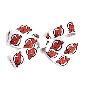 May include: White hair bow featuring a repeating pattern of the New Jersey Devils logo in red and black. The bow is tied in the center with a white knot, and the ribbon is made of a sturdy material.