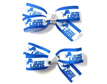 May include: Two white and blue Detroit Lions hair bows. Each bow features the team's logo and name in blue on a white background. The bows are tied with a white center and are set against a white background.