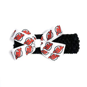May include: A black headband with a white bow featuring a repeating red and white logo. The bow is centered on the headband, creating a decorative accessory. The logo appears to be a stylized letter "N" with a devil's tail.