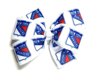 Puede incluir: Lazo blanco para el pelo con el logotipo de los New York Rangers. El lazo está hecho de cinta blanca con el logotipo del equipo impreso repetidamente. El logotipo es un escudo con las palabras "New York Rangers" y una combinación de colores rojo y azul.