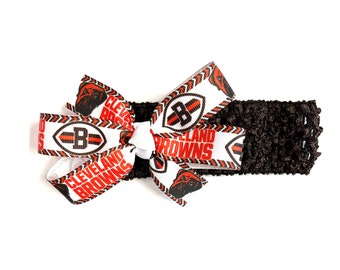 May include: A brown crocheted headband with a white and brown bow. The bow features the words "Cleveland Browns" and the team logo. The headband is a dark brown color.