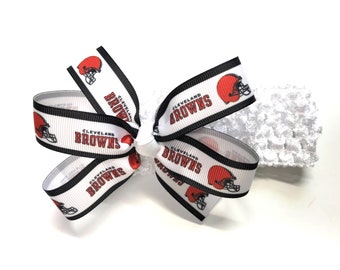 May include: A white hair bow featuring the Cleveland Browns football team logo. The bow has a repeating pattern of the team's helmet and name in brown and black. The bow is attached to a white, textured headband.