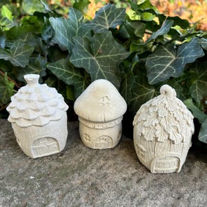 May include: Three miniature fairy houses made of concrete. The houses are shaped like a mushroom, an acorn, and a leaf-covered cottage. The houses are painted in a light gray color.