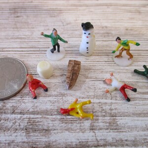 May include: A set of miniature plastic figurines depicting winter activities, including a snowman, a person ice skating, a person skiing, and a person falling down. The figurines are painted in bright colors, including red, green, yellow, and white.