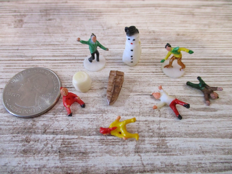 May include: A set of miniature plastic figurines depicting winter activities, including a snowman, a person ice skating, a person skiing, and a person falling down. The figurines are painted in bright colors, including red, green, yellow, and white.