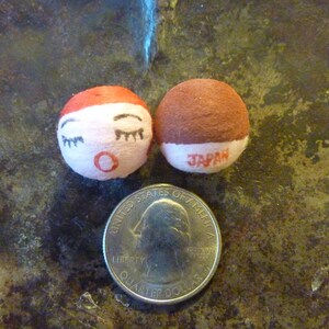 May include: Two small, round Japanese dolls with painted faces and the word "JAPAN" printed on the base. One doll has a red top and the other has a brown top. A U.S. quarter is next to the dolls for size comparison.