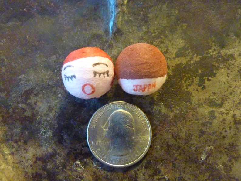 May include: Two small, round Japanese dolls with painted faces and the word "JAPAN" printed on the base. One doll has a red top and the other has a brown top. A U.S. quarter is next to the dolls for size comparison.