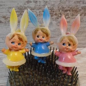 May include: Three small plastic figurines of children wearing bunny ears and dresses. The children are wearing yellow, blue, and pink dresses. The figurines are standing on a bed of tacks.