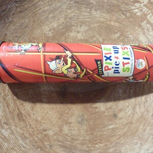 May include: A cylindrical container of Pixie Stix candy. The red container features cartoon characters and the words "Pixie Stix" in a colorful font. The container has a silver metal lid and is set against a wooden background.