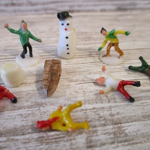 May include: A set of miniature plastic figurines depicting a snowman, a log, and several people in winter clothing. The figurines are white, green, red, yellow, and brown.