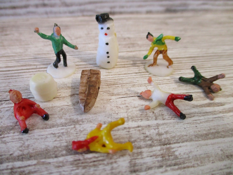 May include: A set of miniature plastic figurines depicting a snowman, a log, and several people in winter clothing. The figurines are white, green, red, yellow, and brown.