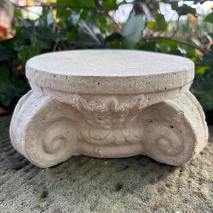 May include: A small, light gray concrete pedestal with a round top and ornate scrollwork on the sides. The pedestal is designed to resemble a classical column capital. The pedestal is sitting on a rough, gray stone surface.