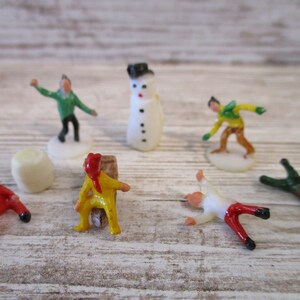 May include: A set of seven miniature plastic figurines, including a snowman, a person in a green shirt, a person in a yellow shirt, a person in a white shirt, a person in a brown shirt, and two people in red shirts. The figurines are on a white wooden surface.