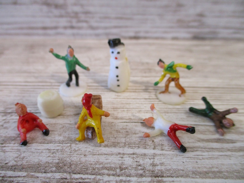 May include: A set of seven miniature plastic figurines, including a snowman, a person in a green shirt, a person in a yellow shirt, a person in a white shirt, a person in a brown shirt, and two people in red shirts. The figurines are on a white wooden surface.