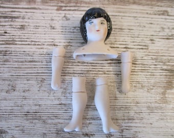 May include: A porcelain doll in pieces, including the head, torso, arms, and legs. The doll has black hair and painted facial features.