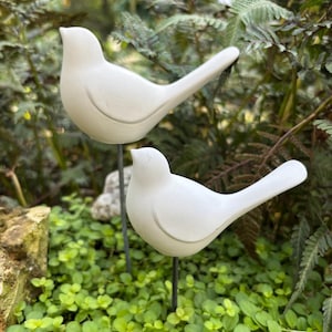 May include: Two white bird garden stakes. The birds are perched on black metal rods, set amidst green foliage and small plants. The birds have a smooth, matte finish and are facing left.