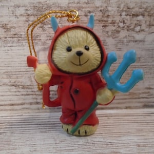 May include: A devil-themed teddy bear ornament. The bear wears a red hooded outfit with blue horns and holds a blue trident. A gold string is attached for hanging. The ornament is on a light wood-grain surface.