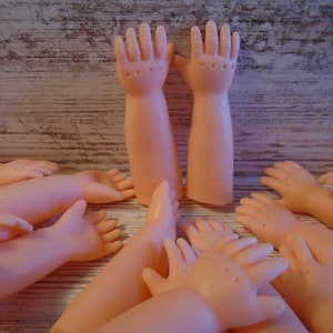 May include: A collection of ten pink plastic doll arms and hands. The arms are all bent at the elbow and the hands are open with fingers spread.