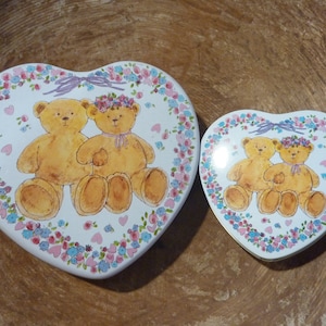 May include: Two heart-shaped tin boxes with a white background and a floral border. The boxes feature a pair of teddy bears wearing floral crowns and holding hands. The larger box is slightly larger than the smaller box.