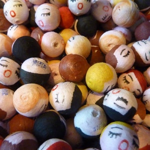 May include: A collection of small, round, hand-painted Japanese dolls. The dolls have closed eyes, red lips, and various hair colors, including black, brown, and yellow. Some dolls have the word "JAPAN" printed on them. The dolls are a mix of colors and textures.