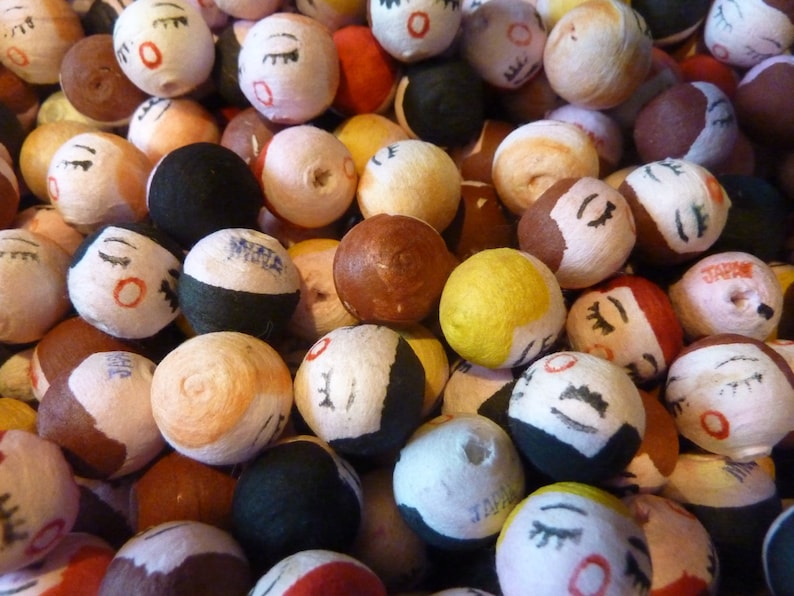 May include: A collection of small, round, hand-painted Japanese dolls. The dolls have closed eyes, red lips, and various hair colors, including black, brown, and yellow. Some dolls have the word "JAPAN" printed on them. The dolls are a mix of colors and textures.