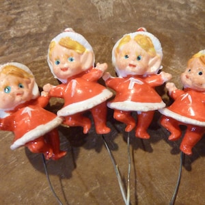 May include: Four vintage Christmas elf figurines. Each elf is wearing a red outfit with white trim and a white hat. The elves have blonde hair and blue eyes. They are attached to thin wire stems.