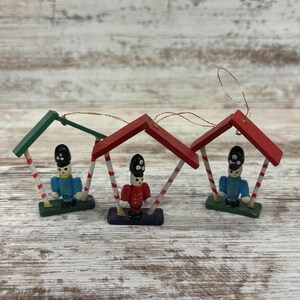 May include: Three wooden toy soldier ornaments. Each soldier is under a red or green roof with white and red striped poles. The soldiers wear black hats and either red or blue jackets. The ornaments have gold string hangers.