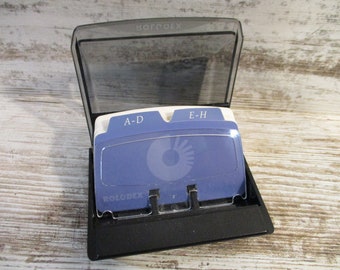 May include: A black Rolodex card file with a clear lid. The file contains blue cards with white lettering and a circular design. The top of the file reads "ROLODEX". The dividers are labeled "A-D" and "E-H".