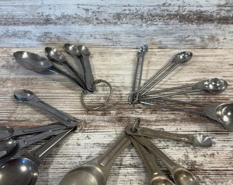 May include: A collection of vintage metal measuring spoons on a wooden surface. The spoons are attached to metal rings and vary in size. The spoons have a worn, antique appearance, suggesting they have been used for many years.