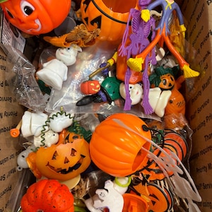 May include: A box filled with various Halloween decorations. The assortment includes orange pumpkin buckets, a purple skeleton, a scarecrow figure, and several small pumpkin and ghost figurines. The items are in a cardboard box.